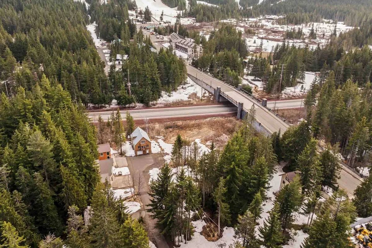 A panoramic scene of dense evergreen forest, snow-covered ground, and distant mountain vistas, with small cabins dotting the landscape. The scenic road hints at nearby towns and adventure beyond the chalet.