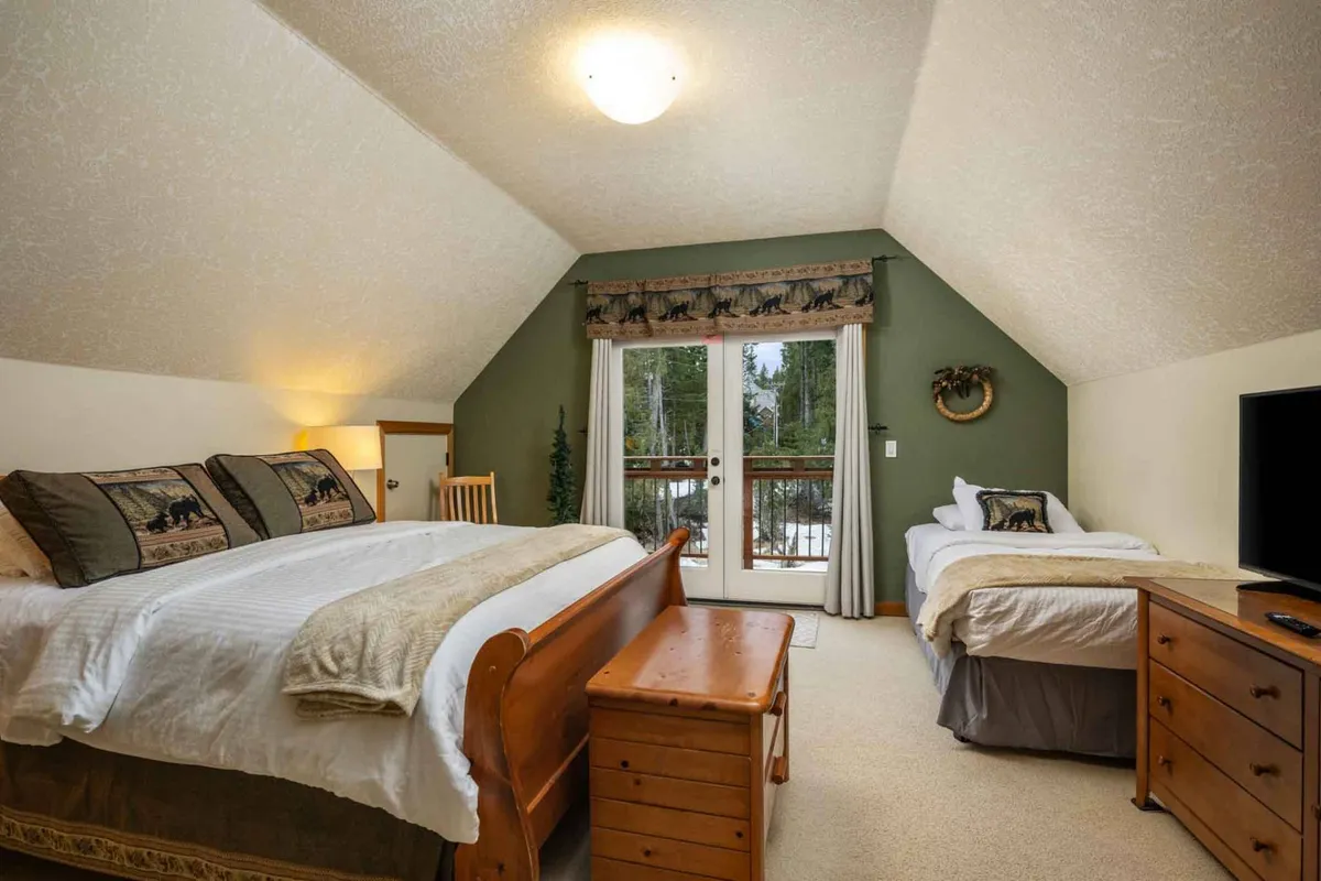 A spacious bedroom featuring a large bed adorned with decorative pillows, double doors opening to a private balcony, a wooden chest at the foot of the bed, and a TV on a dresser. The room is bright, calm, and offers a serene view outdoors.
