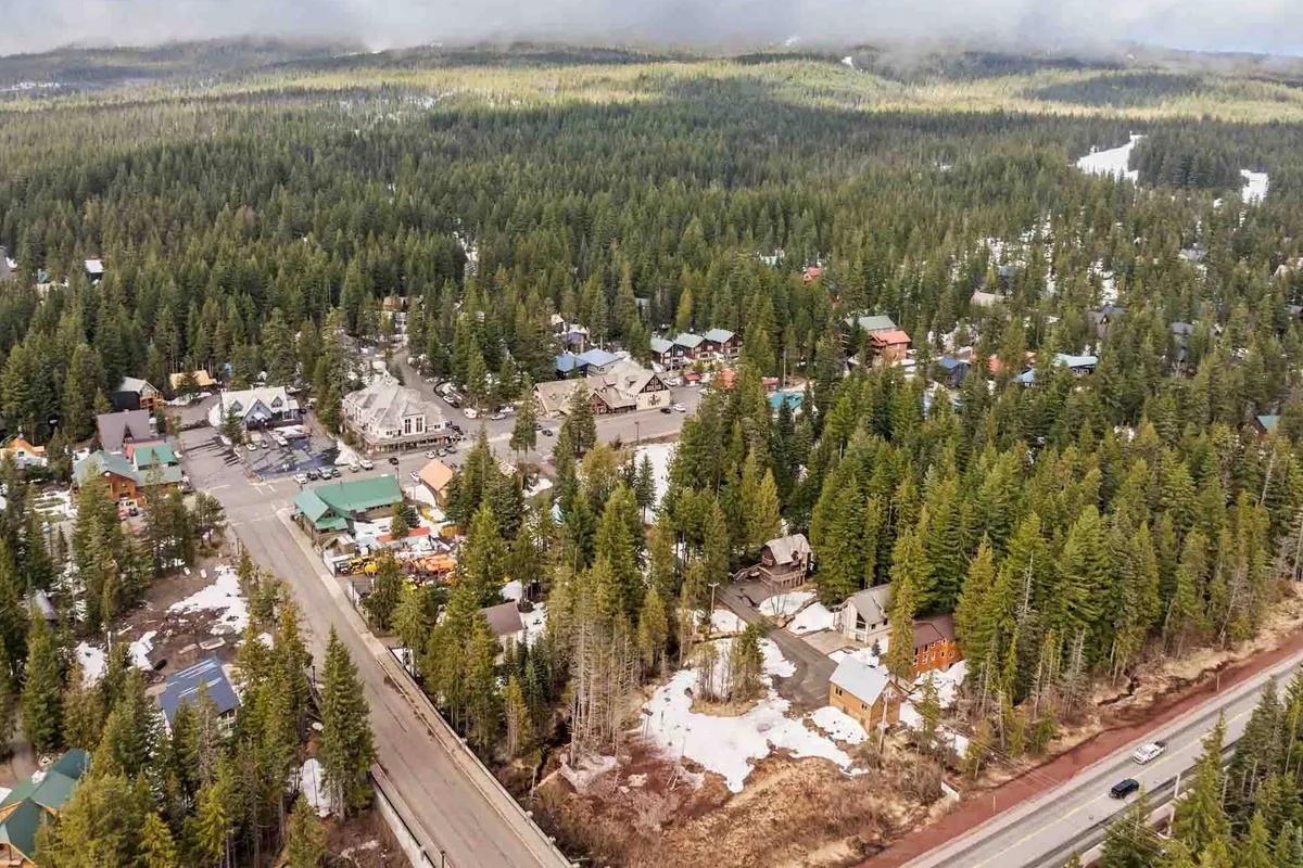 Wide view of a peaceful mountain neighborhood with dense forest, scattered houses, and visible snow patches. The main road invites exploration, while the serene surroundings evoke a sense of community and adventure.