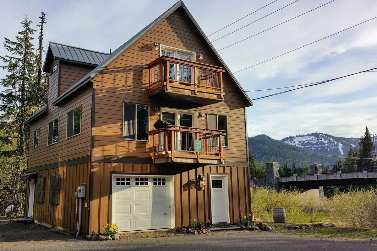 The image features the impressive exterior of Alltitude Chalet, highlighting two inviting balconies perfect for soaking in the mountain scenery, a welcoming front entrance, and views of the surrounding peaks. The scene is set during golden hour, casting a warm glow and emphasizing the cozy, welcoming atmosphere. Guests can imagine relaxing on the balconies, savoring sunset views or a morning coffee while taking in breathtaking mountain vistas.