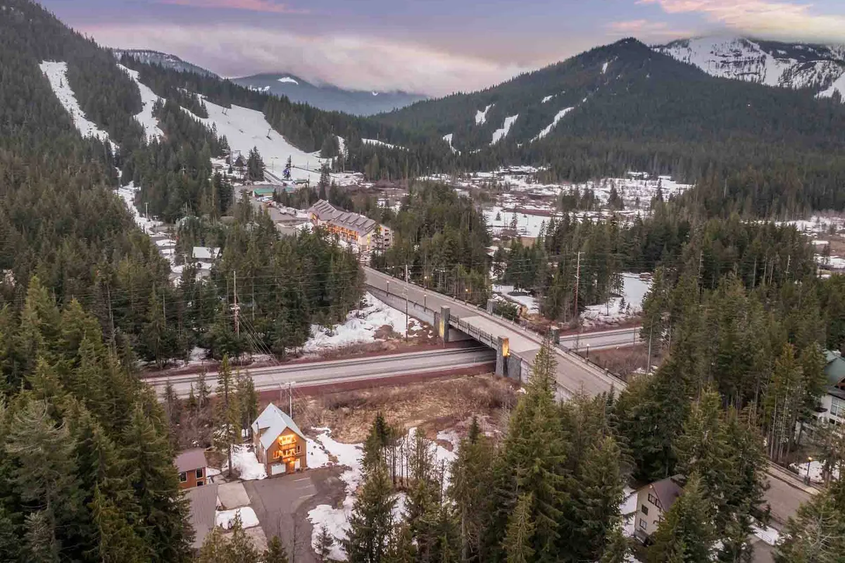 A wide landscape view of snow-covered mountains and dense evergreen forests, with a small, warmly lit house nestled in the scenery and a highway stretching toward the horizon. The composition evokes a sense of connection to nature and opportunity for outdoor exploration.