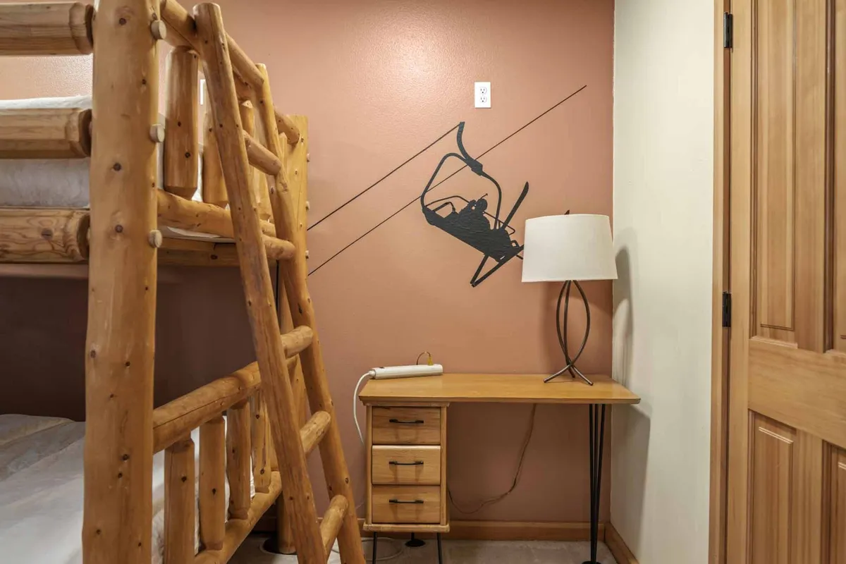 A cozy bunk room featuring a desk with a lamp under wall art depicting a ski lift. The workspace is set up for productivity, and the relaxed atmosphere makes it a versatile space for both work and play. The presence of a bunk bed provides additional flexibility for relaxation or accommodating guests.