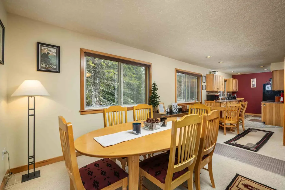 A bright dining area with a large table, ample seating, stylish lamps, and expansive windows. The adjoining kitchen is visible, making this the perfect setting for shared meals, game nights, or festive gatherings with loved ones.
