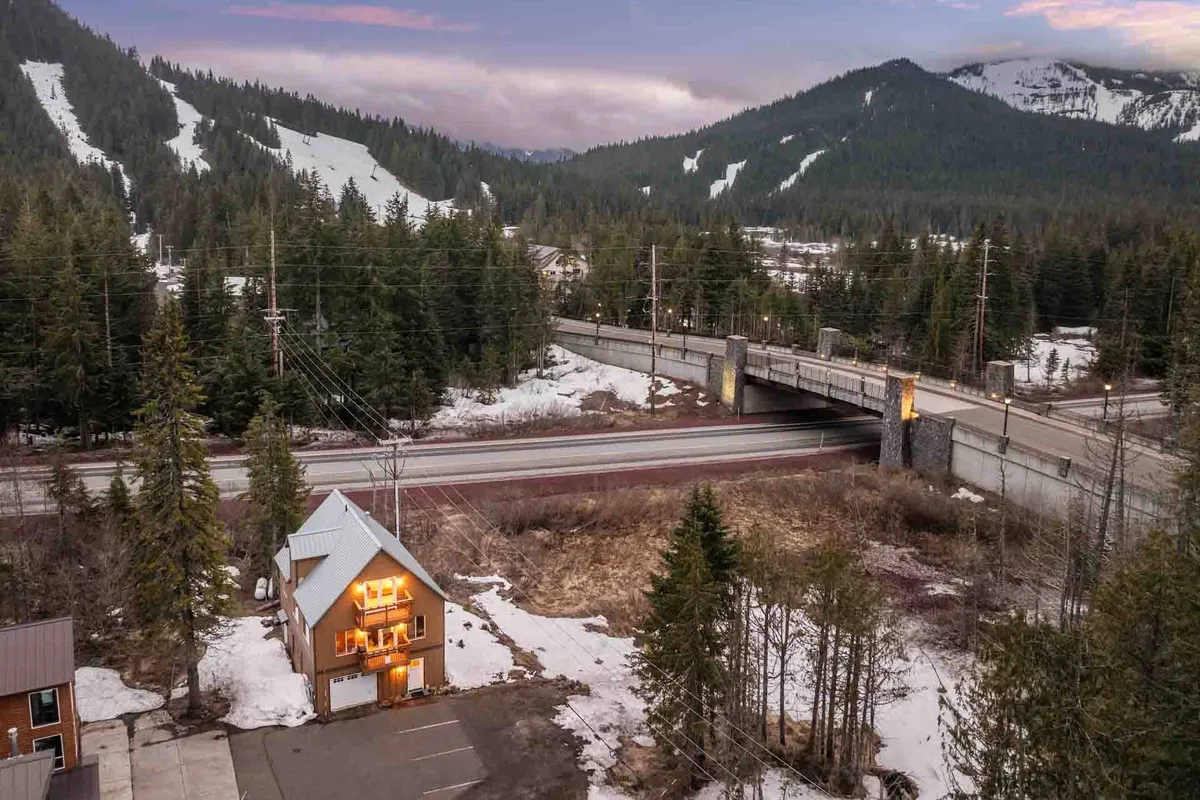 A picturesque exterior view showcasing the cabin nestled against a breathtaking mountain backdrop. Snow blankets the surrounding slopes and a charming bridge leads toward the property, evoking a sense of adventure and tranquility. The visual hints at both winter and summer activities, with the mountain setting inviting for skiing, hiking, and outdoor exploration.
