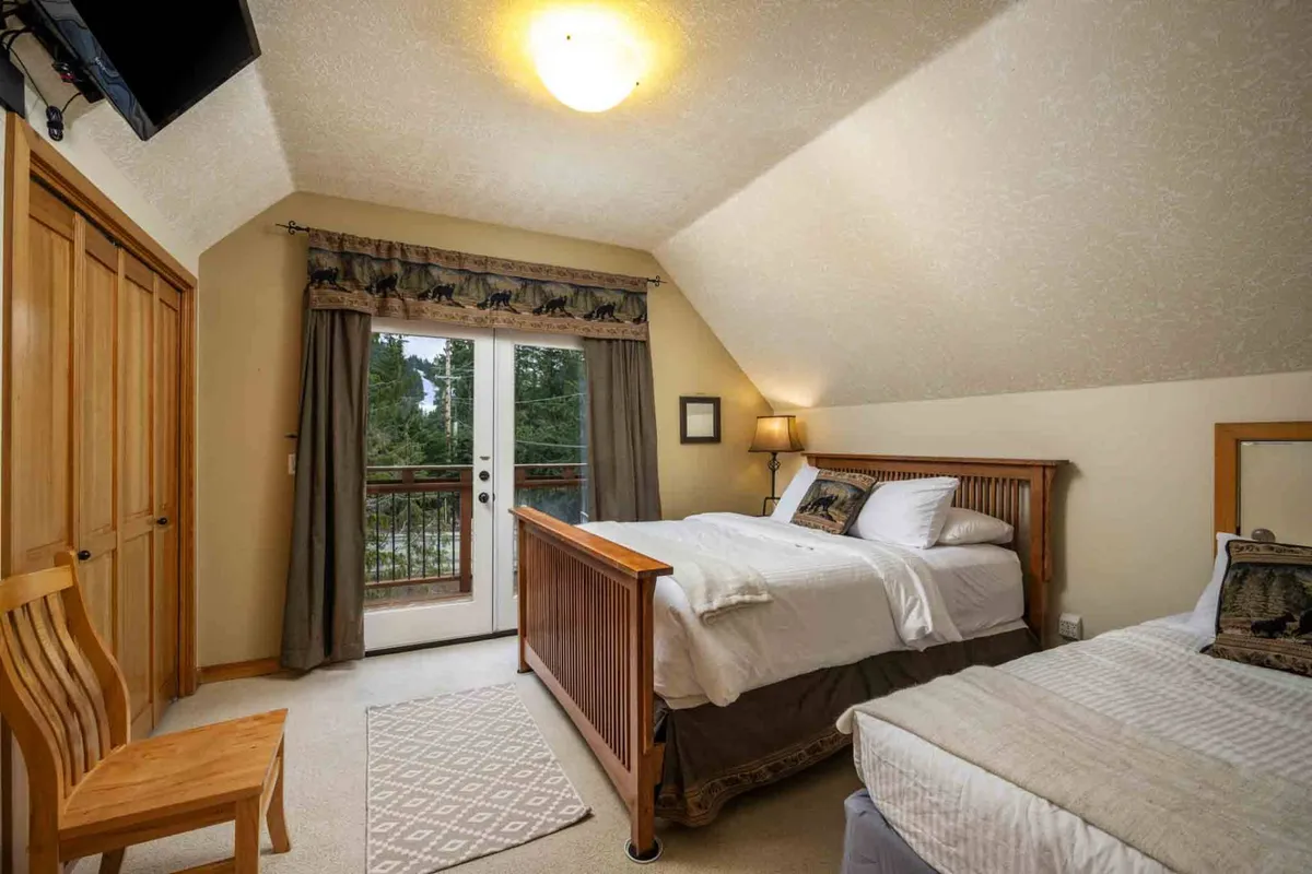 A bright bedroom with two beds and doors opening onto a private balcony. Decorative curtains frame the doors, and morning sunlight streams in, hinting at mountain views just beyond. The space feels calm, inviting, and well-suited for rest or a peaceful work break.